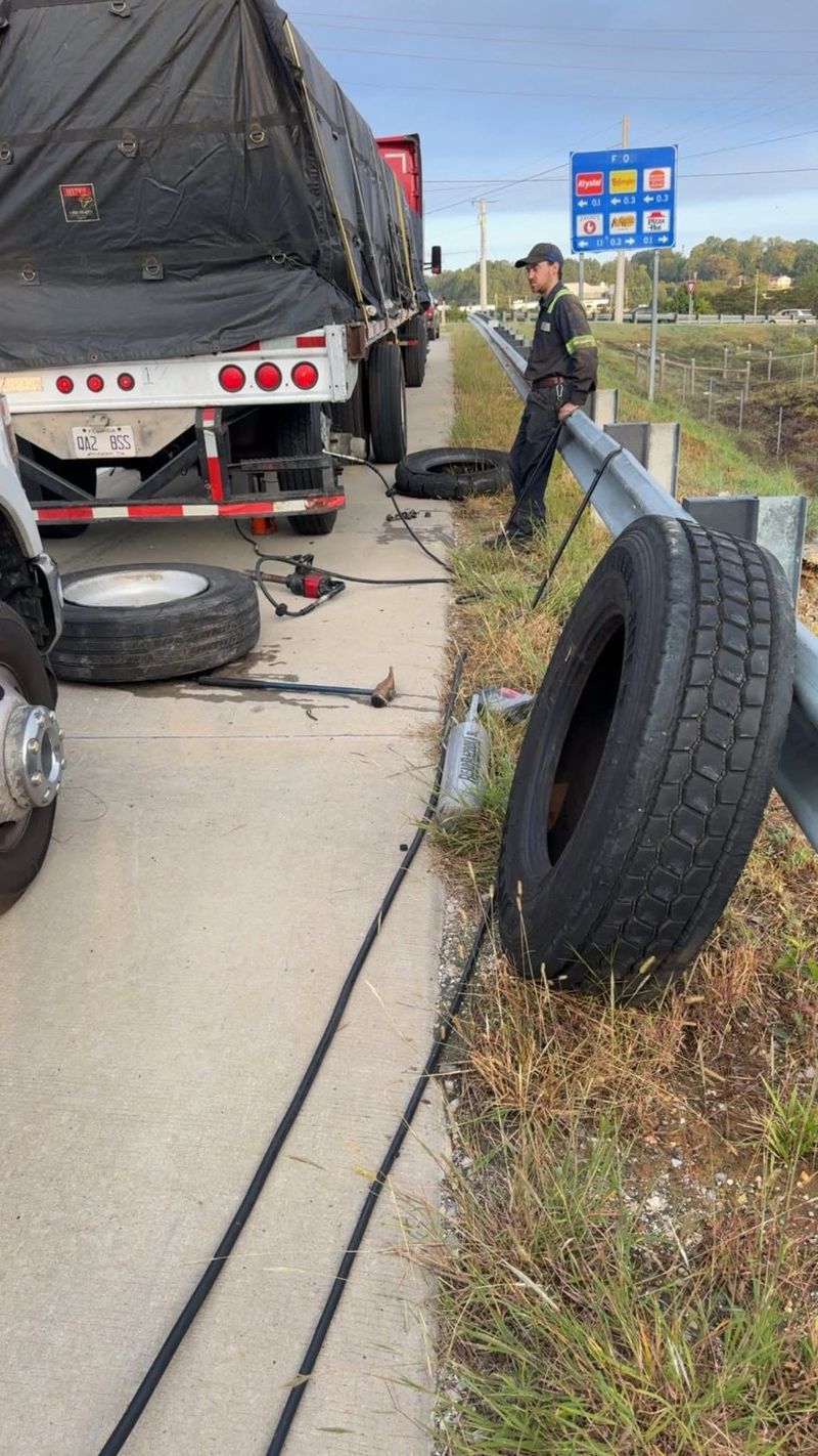 A technician, tires and tools beside a flatbed trailer next to a highway guardrail.
